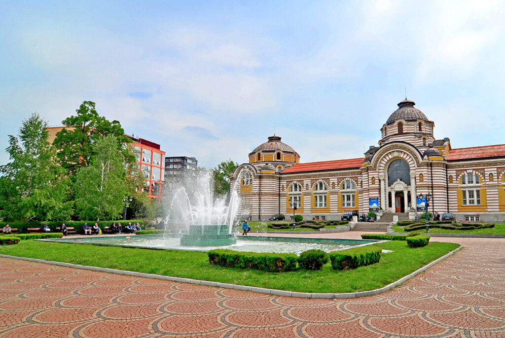 Historic Central Mineral Baths building in Sofia, Bulgaria, with a large fountain and landscaped gardens in the foreground.