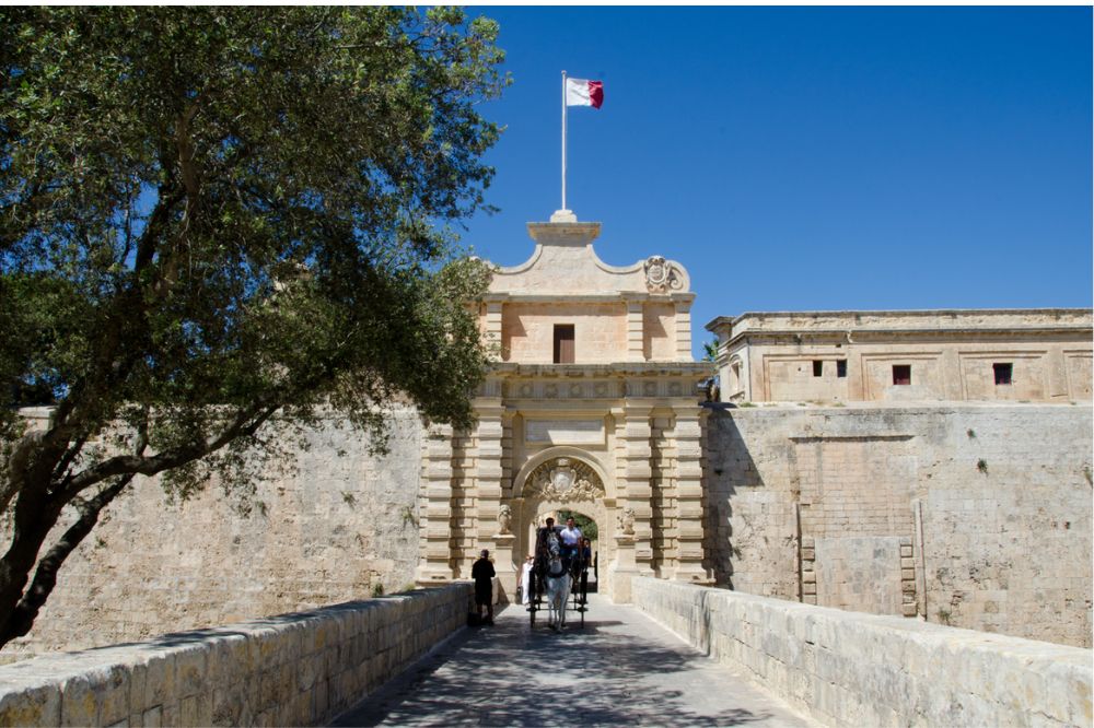 Historic gate of Mdina with a flag overhead, visitors passing through, and a tree casting shade over the bridge.