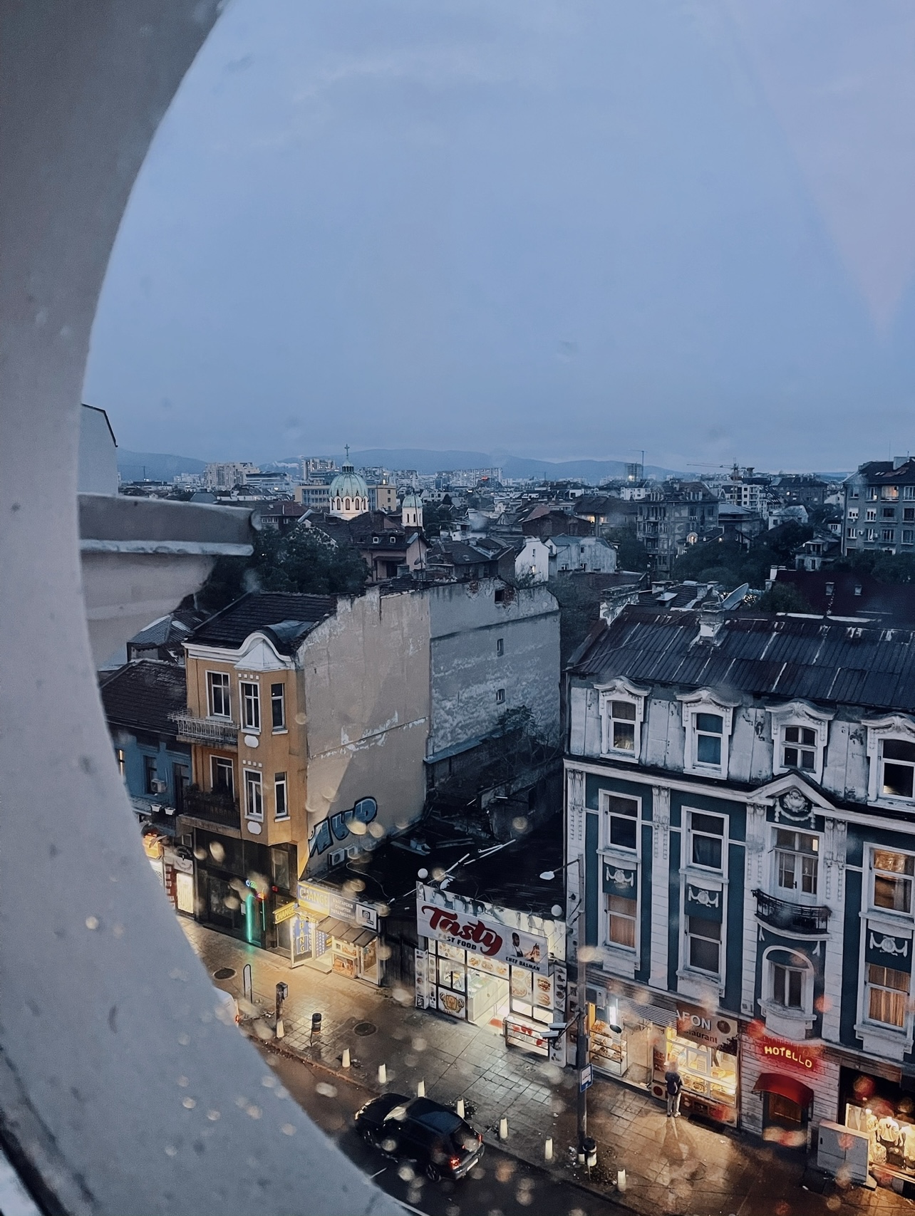 Rain-dappled window view of Sofia at dusk, showing old buildings, lit shopfronts and the city skyline stretching towards distant mountains.