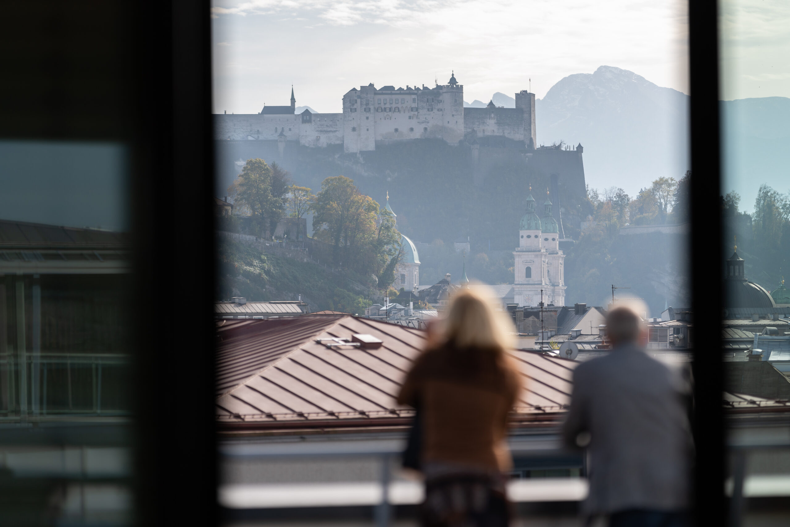 A couple standing on a viewpoint outside Salzburg, overlooking Alpine mountains and valley at golden hour.