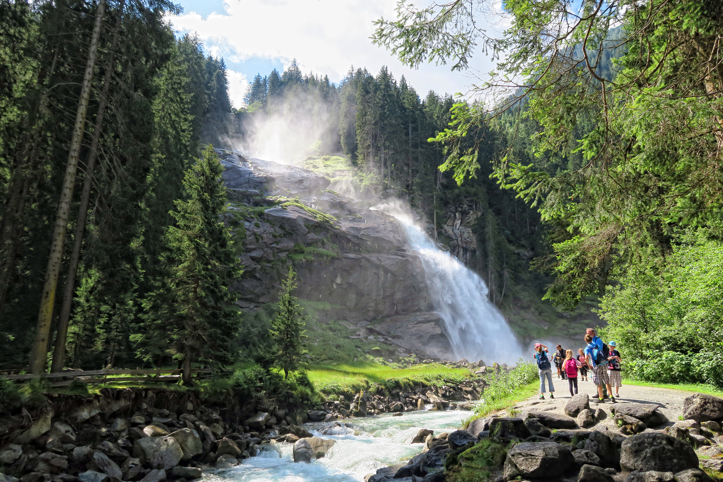 Visitors standing on a viewing platform watching a massive, misty waterfall cascading down steep alpine rock under soft winter light.
