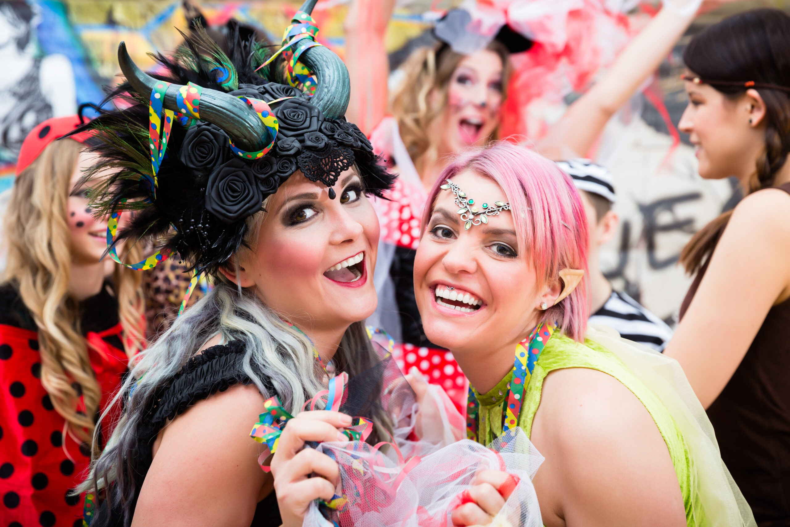 Two smiling women in colourful carnival costumes and makeup, celebrating at a street festival.