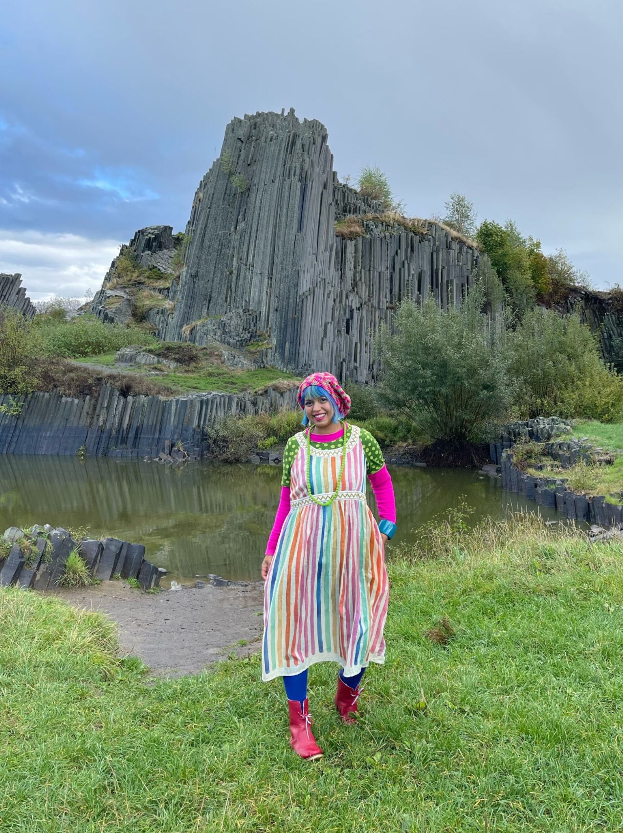 Person in a colorful striped dress and red boots standing in front of the hexagonal basalt columns of Panská Skála in Czechia’s Crystal Valley, surrounded by grass, trees, and a small pond.