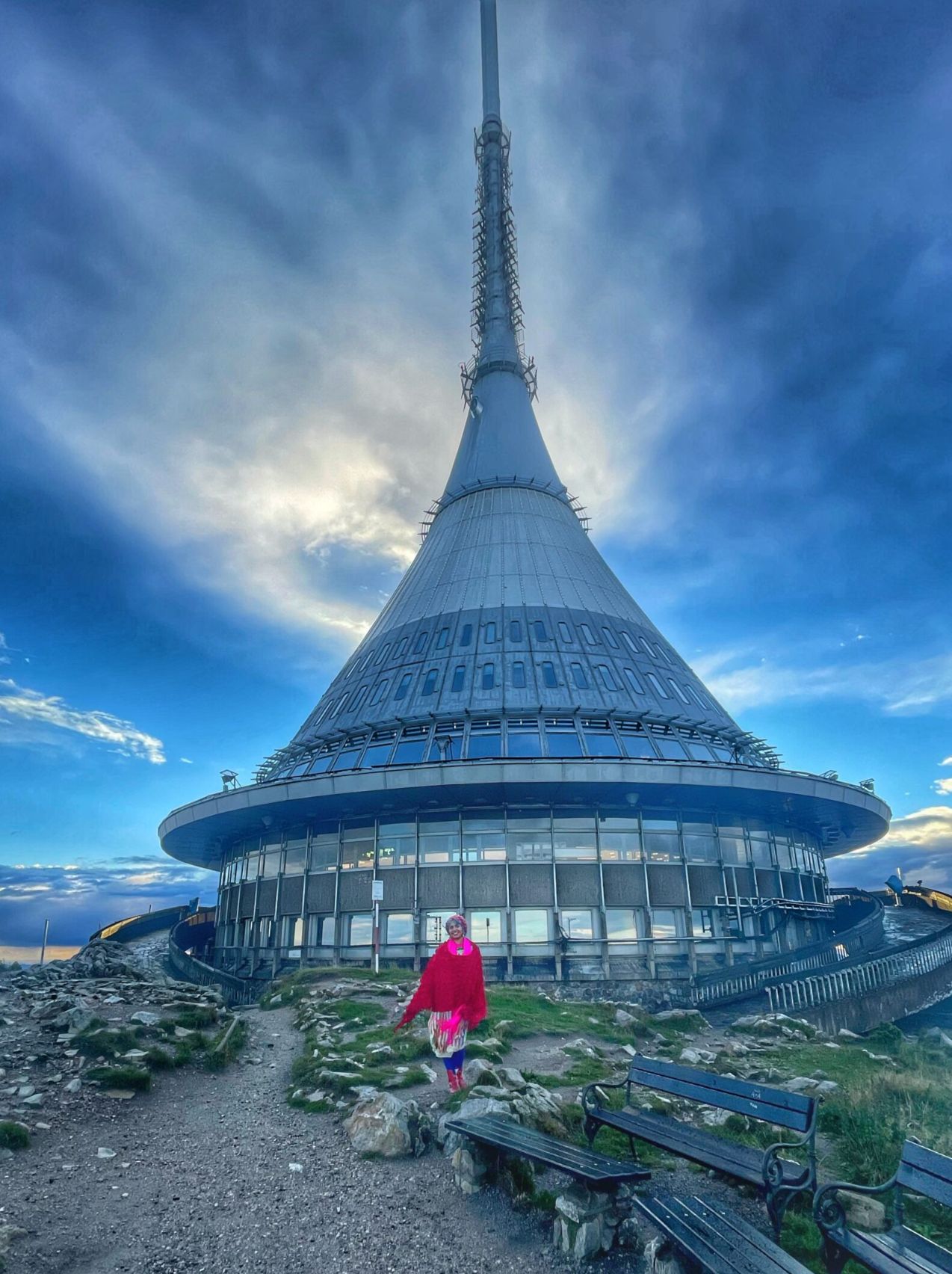A woman wearing a bright pink jacket and scarf stands on a rocky path in front of the futuristic, conical Ještěd Tower at the summit of a mountain, with benches nearby and a dramatic sky overhead.