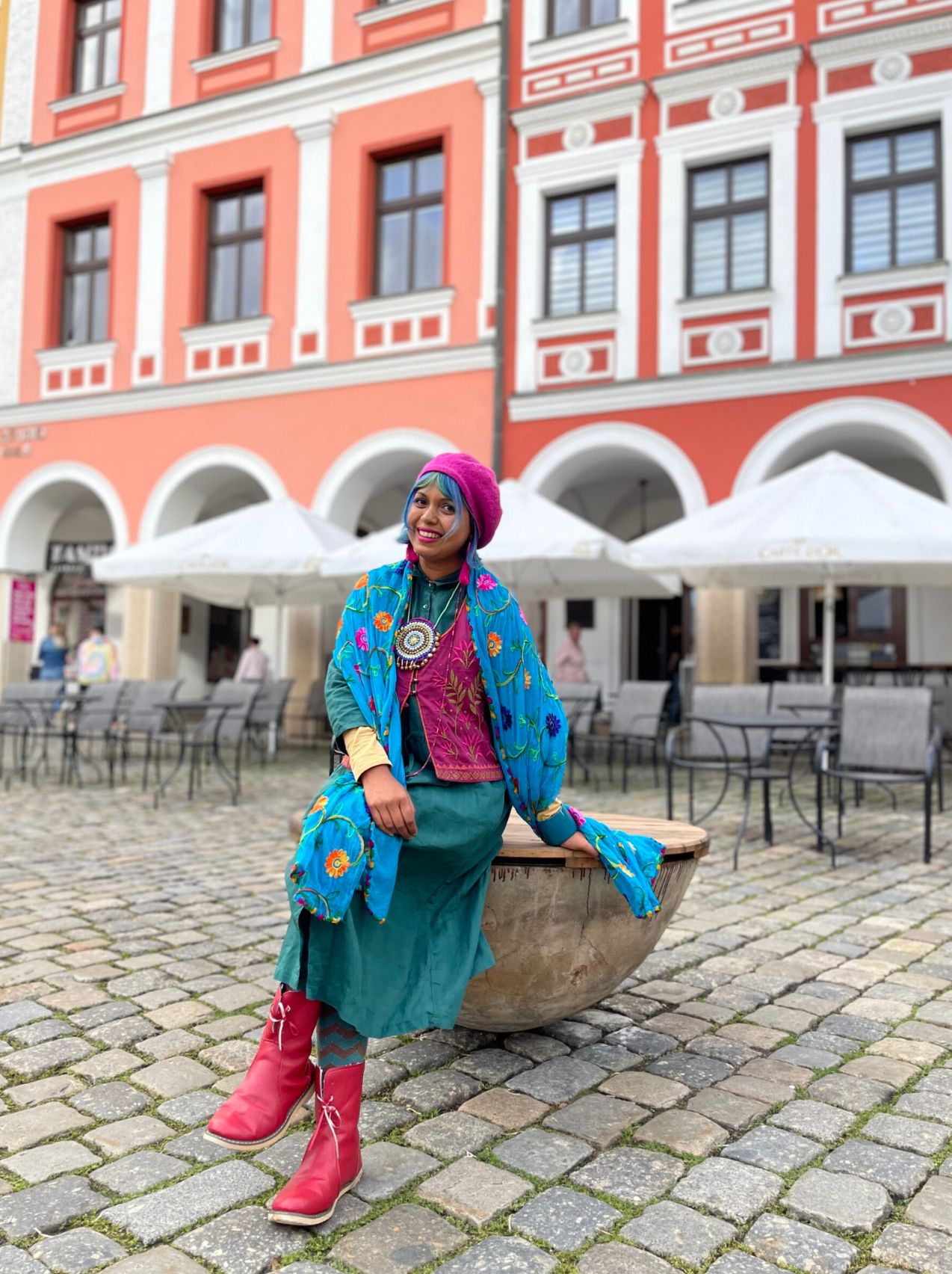 cityscape with historic buildings and rooftops under a soft sky — a woman sits overlooking the city, with urban rooftops and spires extending around her.