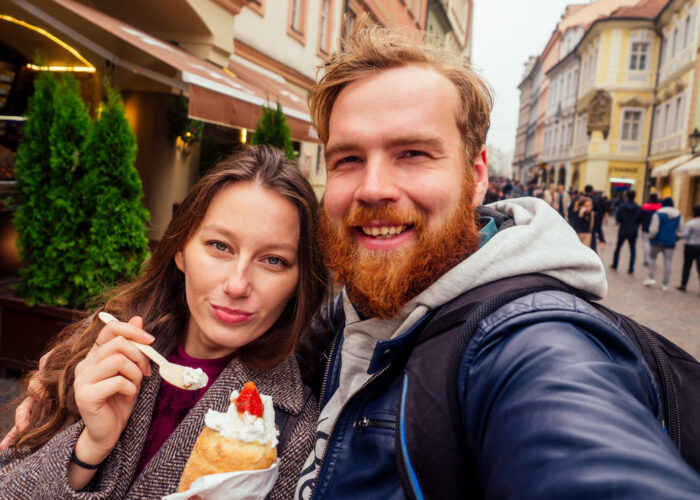 woman and man eating appetite eats a traditional Czech sweet Trdelnik with vanilla cream and strawberries ,useing eco wooden spoon in Prague street.making selfie photo on camera phone.