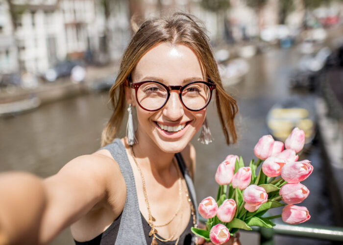 Young beautiful woman making selfie photo with bouguet of pink tulips on the bridge in Amsterdam old city