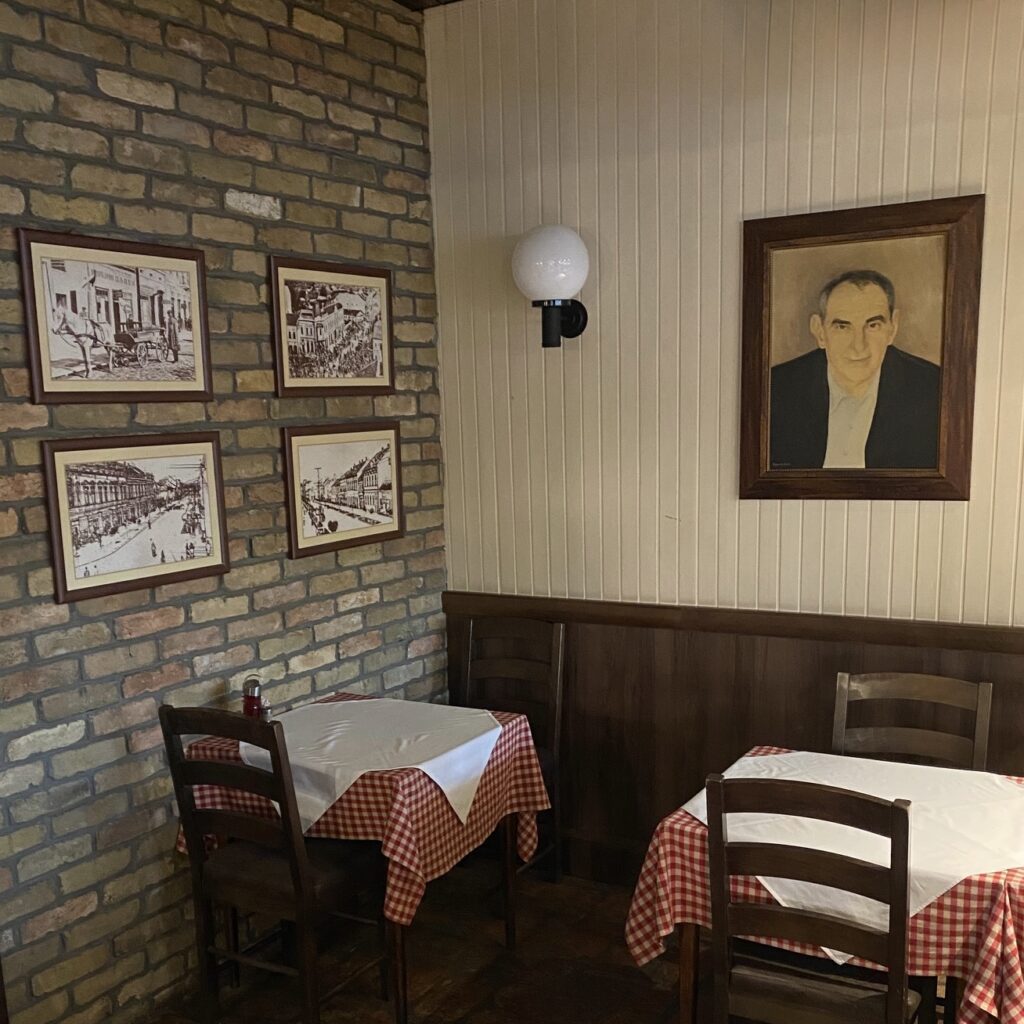 Interior of Šabački Slatinac kafana in Belgrade with checkered tablecloths, wooden chairs, brick walls, and framed photos.