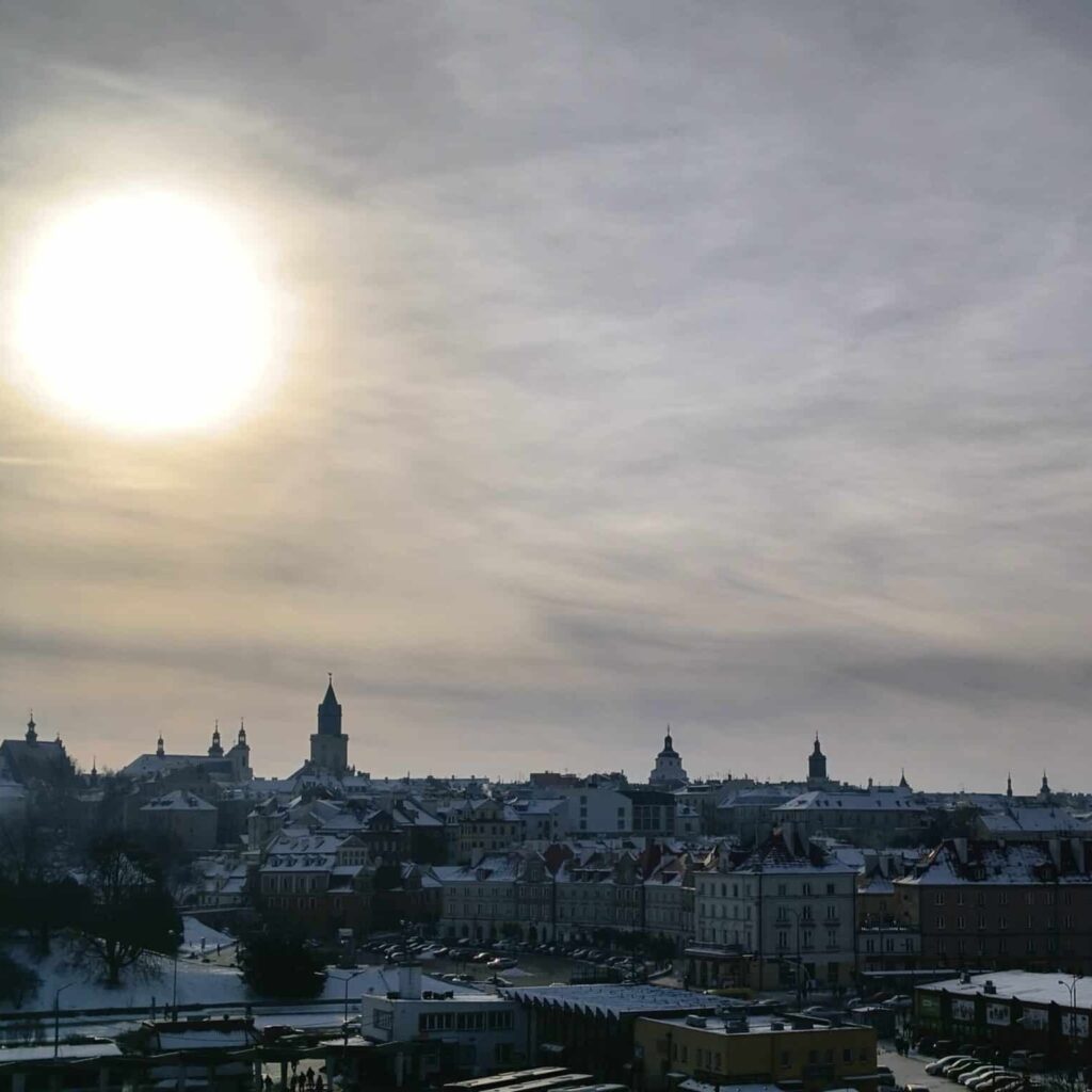 Snow-covered rooftops and church towers across Lublin viewed from Czwartek Hill beneath a hazy winter sun.